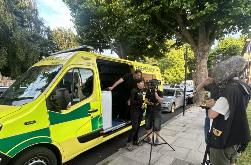 Film crew preparing camera equipment beside an ambulance used as a prop for a film set scene.